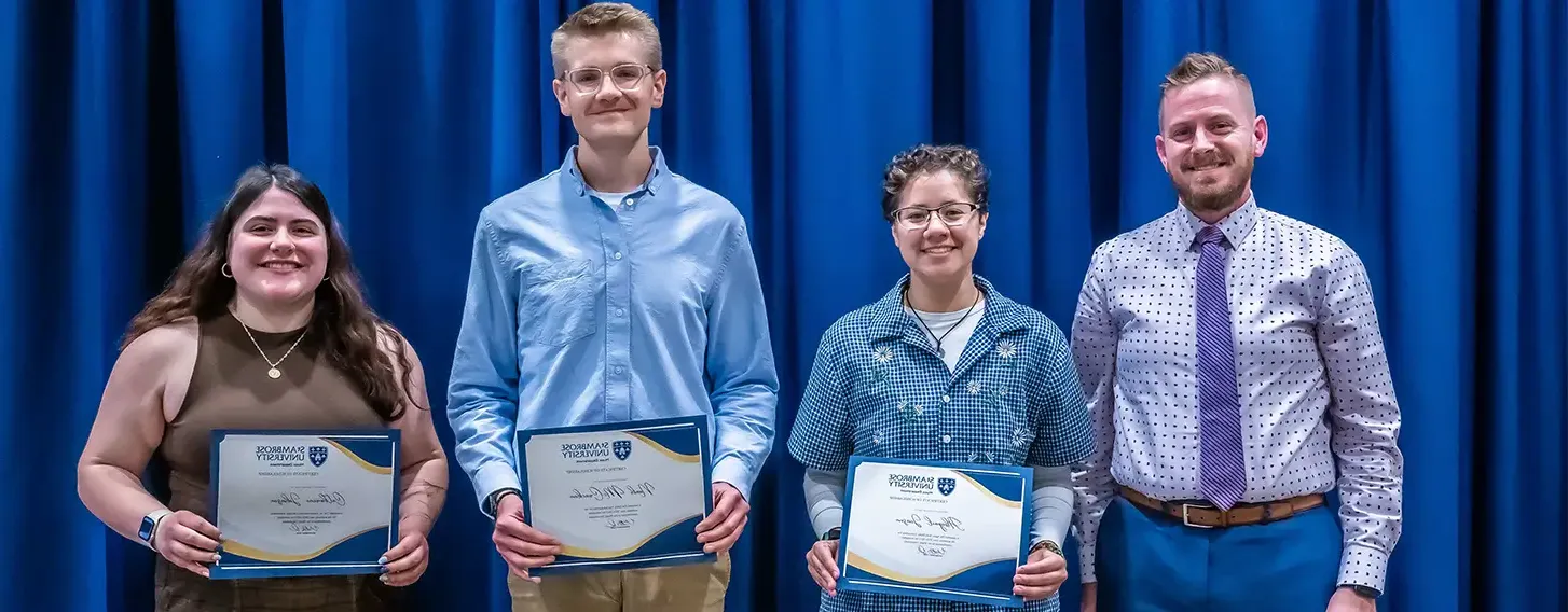 A group of students proudly displaying a scholarship certificate in front of blue curtains.